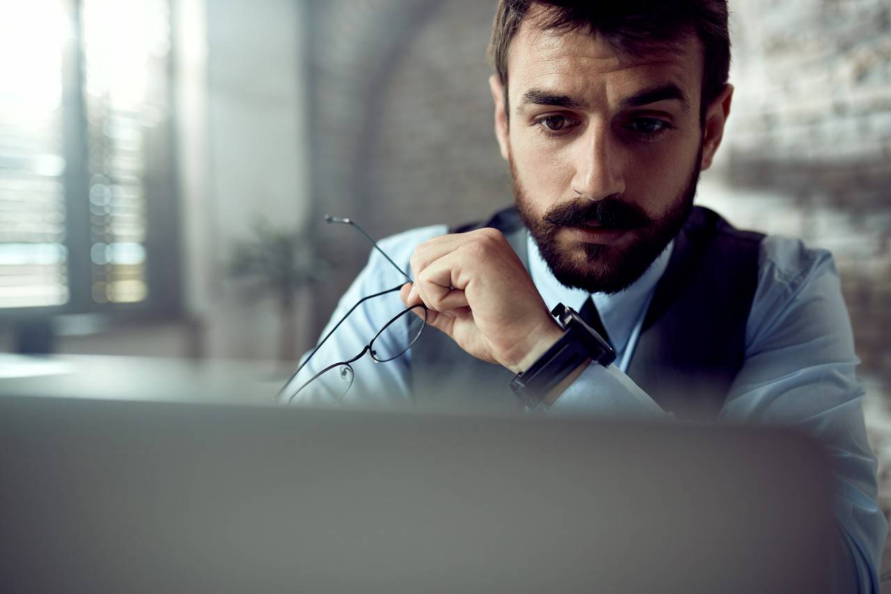 Homem com expressão séria sentado em frente a monitor segurando óculos na mão