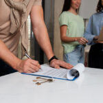Homem assinando contrato em mesa com duas mulheres ao fundo.