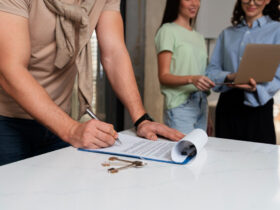 Homem assinando contrato em mesa com duas mulheres ao fundo.