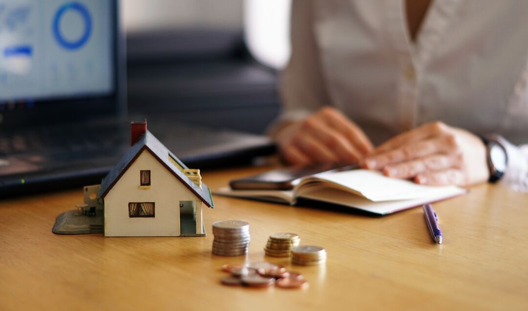 Casa em miniatura com moedas e laptop ao fundo